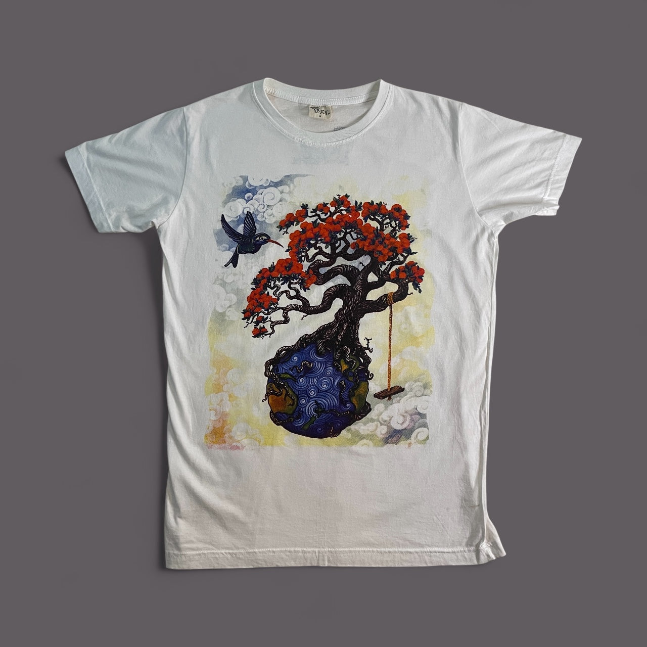Maglietta bianca unisex in cotone con stampa artistica di un albero fiorito radicato in un pianeta blu e un uccello in volo.