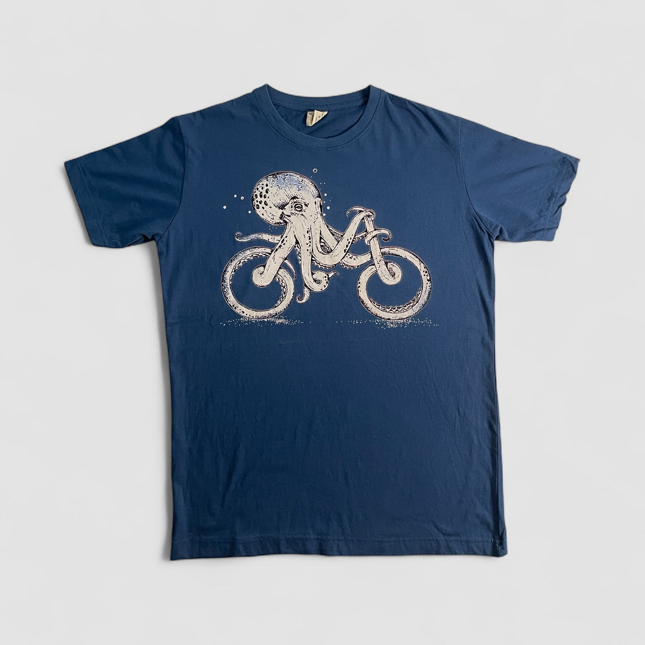 Maglietta blue unisex in cotone con stampa artistica di un polpo che pedala una bicicletta, stile creativo e alternativo.