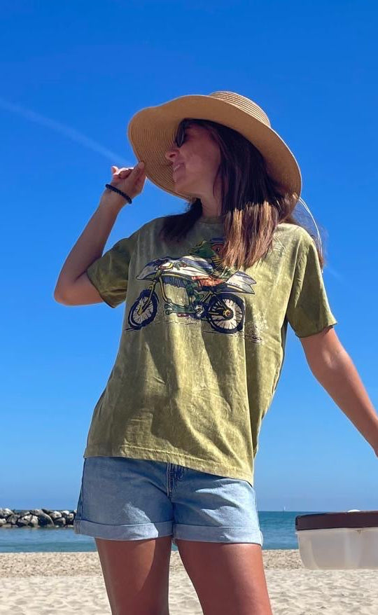 Maglietta verde vintage unisex con stampa rana su bici e tavola da surf “Frog Rider”, indossata, con sfondo spiaggia.